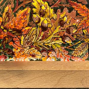 May include: A rectangular artwork featuring a fall-themed design with yellow, orange, and brown leaves, acorns, and berries. The artwork is mounted on a light-colored wooden base, suitable for display. The artwork is likely a decorative piece.