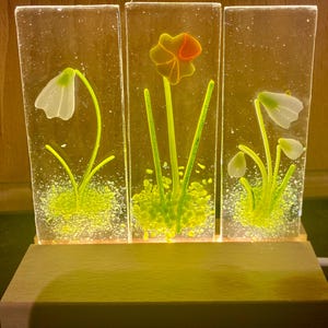 Könnte beinhalten: Dekorative Glasplatte mit drei rechteckigen Abschnitten, die jeweils ein Design aus geschmolzenen Glasblumen aufweisen. Die Blumen umfassen weiße Schneeglöckchen und eine gelbe Narzisse mit orangefarbenen Akzenten, vor einem hellgrünen Hintergrund. Die Platte steht auf einem Holzsockel.