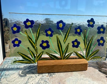 May include: A glass panel with a painted design of blue flowers with yellow centers and green stems. The panel is mounted on a wooden stand.