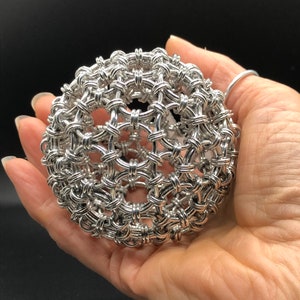 May include: A silver metal sphere made of interwoven wire, creating a geometric pattern. The sphere is being held in a person's hand.