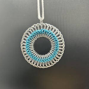 May include: A silver and teal blue chainmail pendant in the shape of a circle. The pendant is hanging from a white cord.