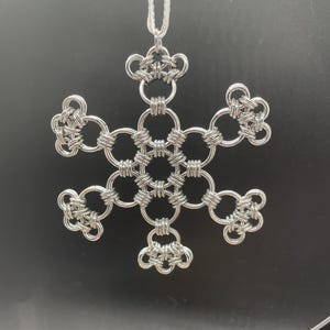 May include: A silver-colored snowflake ornament, intricately crafted from interwoven metal rings. The snowflake design features six symmetrical arms, each with a unique looped pattern. A white cord is attached for hanging. The background is a solid black.