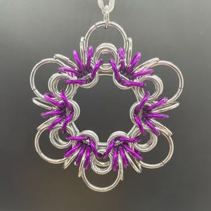 May include: A silver-colored chainmail pendant in a flower shape with five petals. Each petal has a cluster of bright purple chainmail rings at the center. The pendant hangs from a thin, silver-colored chain.