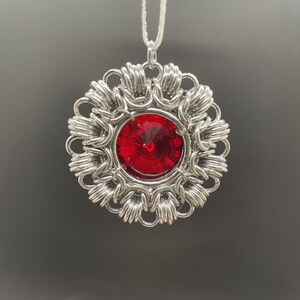 Op de afbeelding: Een zilveren hanger met een rood juweel in het midden, omgeven door een delicaat, ingewikkeld ontwerp. De hanger is bevestigd aan een zilveren ketting.