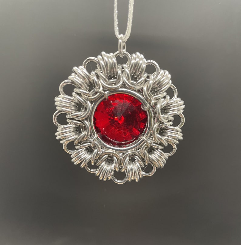 Op de afbeelding: Een zilveren hanger met een rood juweel in het midden, omgeven door een delicaat, ingewikkeld ontwerp. De hanger is bevestigd aan een zilveren ketting.