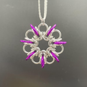 May include: A silver-colored chainmaille pendant with a circular design. The pendant features a central ring surrounded by a series of interlocking rings, with vibrant purple accents. The pendant hangs from a thin, silver-colored chain.