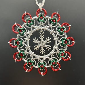 Könnte beinhalten: Ein kreisförmiges Kettenhemd-Ornament aus ineinandergreifenden silbernen, roten und grünen Metallringen. Ein silberfarbener Schneeflocken-Anhänger hängt in der Mitte der Ringstruktur, die an einem dünnen weißen Band befestigt ist.
