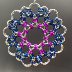 May include: A handcrafted, circular ornament with a silver-colored outer ring and intricate inner detailing. The design incorporates blue and purple rings, creating a visually appealing, symmetrical pattern. A silver string is attached for hanging.