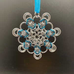 May include: A silver snowflake ornament with blue accents hangs from a blue ribbon. The snowflake is made of interlocking metal rings.