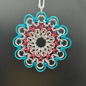 May include: A handmade ornament with a floral design. The ornament is made of interlocking metal rings in teal, silver, and red. It is suspended by a white ribbon. The ornament is circular with a central hole.