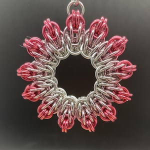May include: A silver and pink metal chainmail pendant in the shape of a flower with a circular opening in the center.