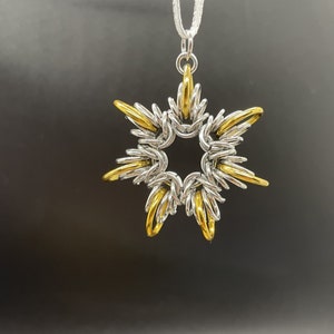 Pode incluir: Um pingente em forma de estrela de prata e ouro com uma corrente presa. O pingente é feito de anéis de metal entrelaçados.