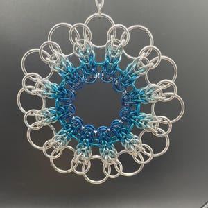 以下が含まれることがあります： 銀色と青色の金属リングで作られた円形のペンダント。デザインは、中央の円と、花や雪の結晶のようなパターンを作り出す一連の相互接続されたリングで構成されています。青色は中央の濃い色合いから明るい色合いに変化します。