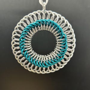 May include: A silver-colored chainmail pendant with a teal-colored inner ring. The pendant is circular and hangs from a silver ribbon. The chainmail is made of small, interlocking metal rings.