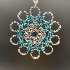 May include: A silver and teal blue snowflake ornament made of metal rings. The ornament is hanging from a white ribbon.