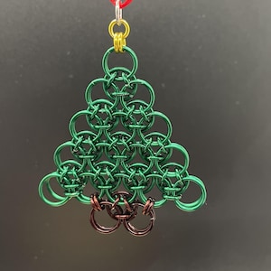 May include: A chainmail ornament shaped like a Christmas tree, constructed from interlocking green metal rings with a brown metal ring base. The ornament features a gold-colored ring at the top, attached to a thin red ribbon for hanging purposes.