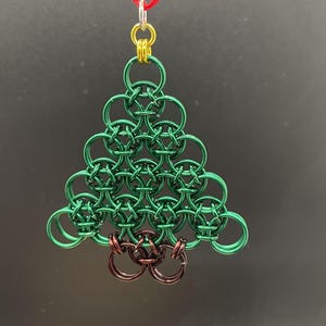 May include: A chainmail ornament shaped like a Christmas tree, constructed from interlocking green metal rings. The base features brown metal rings, while the top is accented with gold rings and a silver jump ring attached to a thin red ribbon.