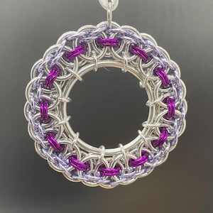 Puede incluir: Adorno circular hecho a mano con un diseño de cota de malla. El adorno presenta una base de color plateado con secciones de anillos morados y magenta. Se adjunta una cinta plateada para colgarlo. El fondo es gris oscuro.