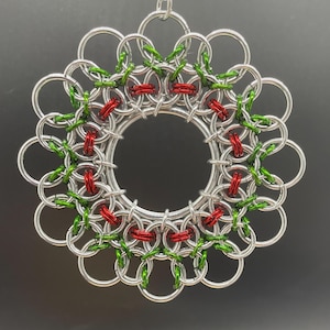 以下が含まれることがあります： 緑と赤のアクセントが施された、円形のデザインの銀色の金属製オーナメント。オーナメントは、お互いに絡み合ったリングでできており、お祝いの装飾品を作り出しています。白いリボンから吊り下げられており、休日の装飾に適しています。