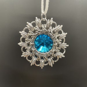 Op de afbeelding: Een zilveren hanger met een blauwe edelsteen in het midden, omringd door een delicaat, ingewikkeld ontwerp. De hanger is bevestigd aan een wit koord.