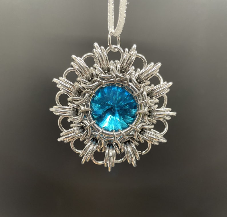 Op de afbeelding: Een zilveren hanger met een blauwe edelsteen in het midden, omringd door een delicaat, ingewikkeld ontwerp. De hanger is bevestigd aan een wit koord.