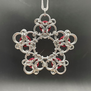 May include: A silver-colored, chainmail-style star-shaped ornament with red accents. The ornament is suspended by a thin, white cord. The design features a central circular opening and five points, each with a series of interlocking rings.