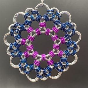May include: A handcrafted, circular ornament with a silver-colored outer ring and intricate inner detailing. The design incorporates blue and purple rings, creating a visually appealing, symmetrical pattern. A silver string is attached for hanging.