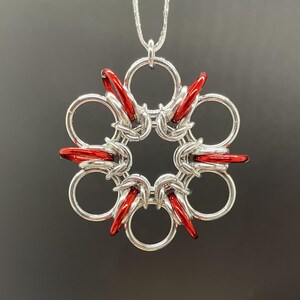Peut inclure: Un collier pendentif argenté avec un motif floral. Le pendentif présente une série d'anneaux interconnectés, avec des accents rouges. Le collier est suspendu à une fine chaîne argentée. Le fond est gris foncé uni.