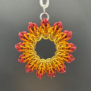 May include: A red and gold metal pendant in the shape of a flower with a circular center. The pendant is hanging from a silver chain.