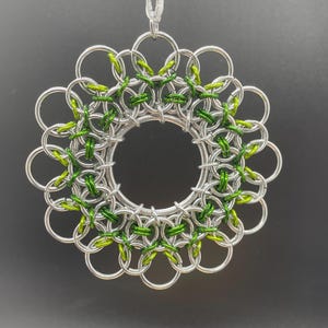May include: A silver-colored chainmail ornament with a floral design. The ornament features a central circle surrounded by interlocking rings, with green accents woven throughout. A white string is attached for hanging.