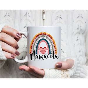 Puede incluir: Taza de cer&aacute;mica blanca con un dise&ntilde;o de arco&iacute;ris y el texto "Mamacita" en letras negras. El arco&iacute;ris est&aacute; formado por rayas rojas, naranjas, amarillas, verdes, azules y moradas. Hay un coraz&oacute;n rosa en el centro del arco&iacute;ris.