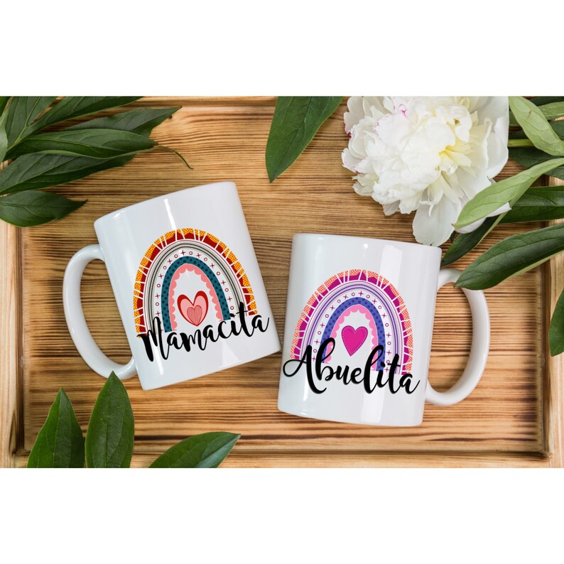 Puede incluir: Dos tazas de cer&aacute;mica blanca con dise&ntilde;os de arco&iacute;ris y corazones. Una taza dice "Mamacita" y la otra dice "Abuelita".