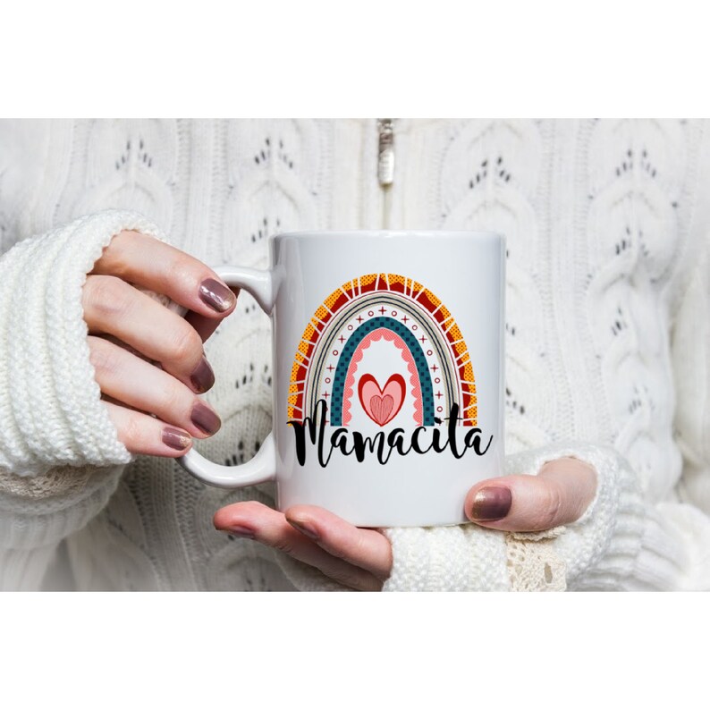 Puede incluir: Taza de cer&aacute;mica blanca con un dise&ntilde;o de arco&iacute;ris y el texto "Mamacita" en letras negras. El arco&iacute;ris est&aacute; formado por rayas rojas, naranjas, amarillas, verdes, azules y moradas. Hay un coraz&oacute;n rosa en el centro del arco&iacute;ris.