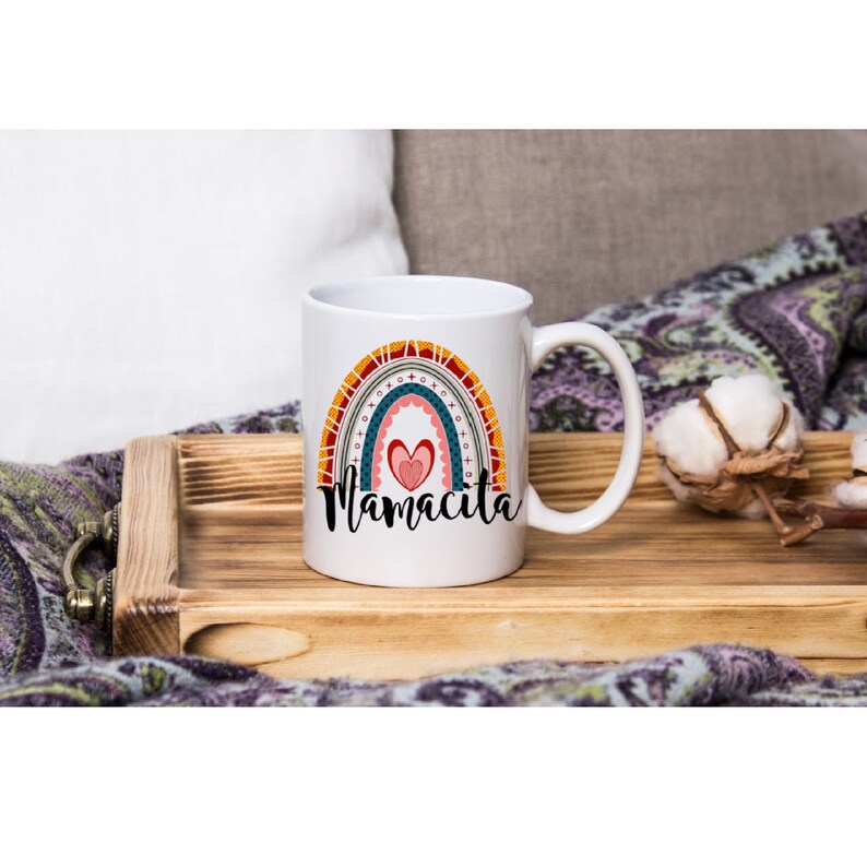 Puede incluir: Taza de cer&aacute;mica blanca con un dise&ntilde;o de arco&iacute;ris y el texto "Mamacita" en negro. El arco&iacute;ris est&aacute; formado por rayas rojas, naranjas, amarillas, verdes, azules y moradas. Hay un coraz&oacute;n rosa en el centro del arco&iacute;ris.