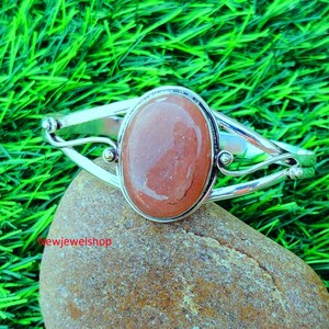 May include: A silver bangle bracelet with a large, oval-shaped, reddish-brown gemstone set in the center. The bracelet has a delicate, ornate design.