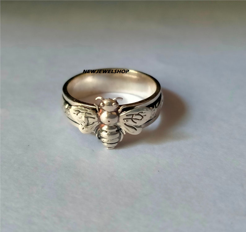 May include: A silver ring featuring a detailed bee design. The bee is positioned centrally on the ring, with wings extending outwards. The ring is a plain band with a subtly textured surface.