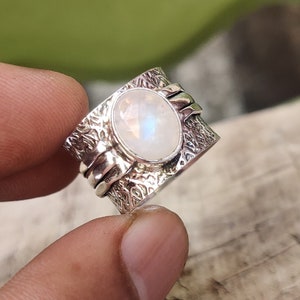 May include: A silver ring featuring a large oval moonstone set in a textured band. The band is wide and has a hammered finish.