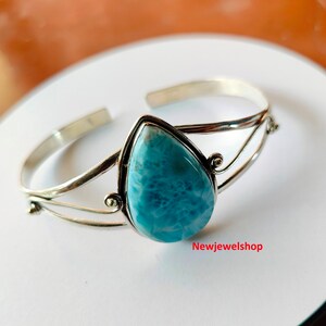 May include: A silver cuff bracelet with a light blue, pear-shaped gemstone. The gemstone is set in a silver bezel, and the bracelet has an open design. The text "Newjewelshop" is visible on the surface.