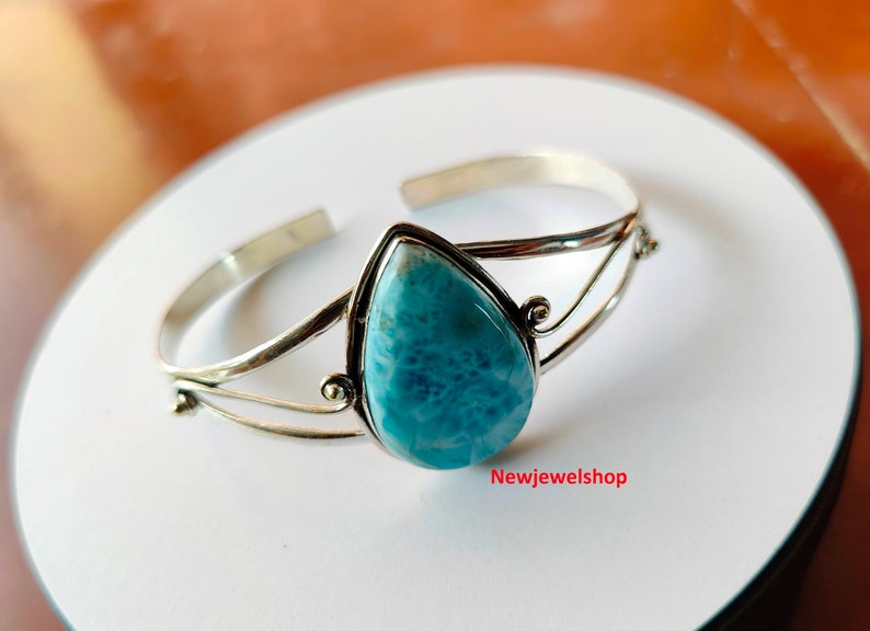 May include: A silver cuff bracelet with a light blue, pear-shaped gemstone. The gemstone is set in a silver bezel, and the bracelet has an open design. The text "Newjewelshop" is visible on the surface.