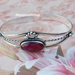 May include: Silver bangle bracelet featuring a red gemstone set in a silver bezel. The bracelet has a twisted wire design and a decorative silver accent.