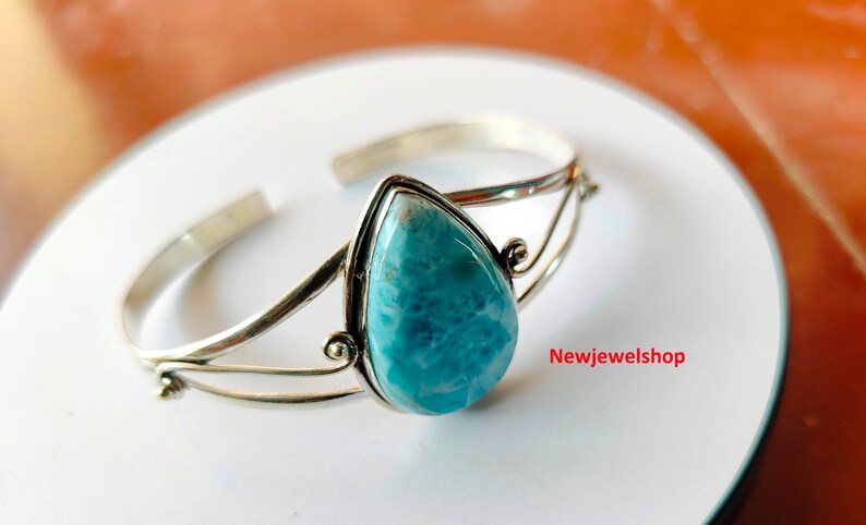 May include: A silver cuff bracelet with a pear-shaped, light blue gemstone. The open design has decorative swirls near the gemstone. The bracelet is displayed on a white surface.