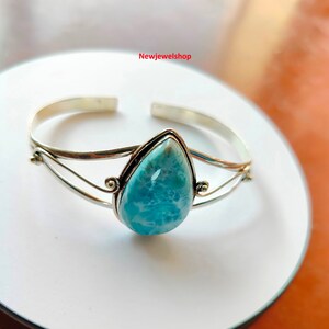 May include: A silver cuff bracelet with a pear-shaped, light blue larimar stone. The stone is set in a silver bezel. The bracelet has a simple, elegant design. The bracelet is displayed on a white surface.