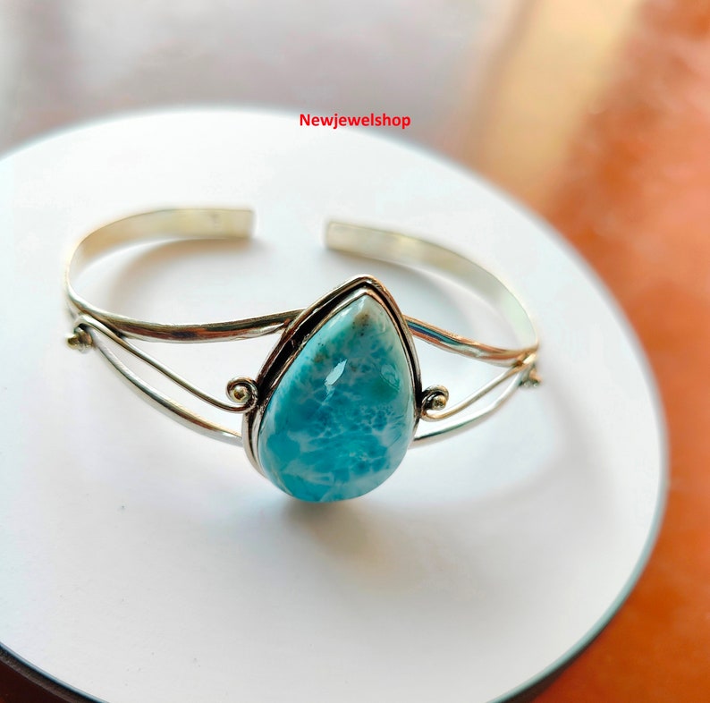 May include: A silver cuff bracelet with a pear-shaped, light blue larimar stone. The stone is set in a silver bezel. The bracelet has a simple, elegant design. The bracelet is displayed on a white surface.