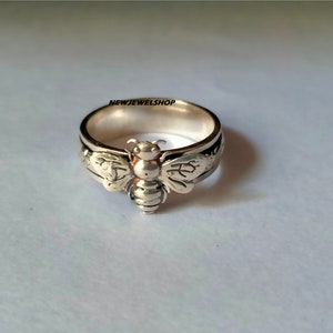 May include: A silver ring featuring a detailed bee design. The bee is positioned centrally on the ring, with wings extending outwards. The ring is a plain band with a subtly textured surface.