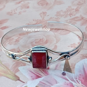 May include: A silver bangle bracelet featuring a red gemstone set in a square shape. The bracelet has a delicate, ornate design with a wavy pattern.