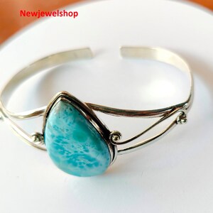 May include: A silver cuff bracelet featuring a pear-shaped, light blue larimar stone. The stone is set in a silver bezel. The bracelet has a simple, elegant design. The jewellery is displayed on a white surface.
