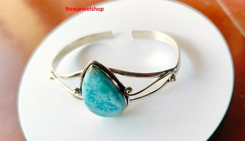 May include: A silver cuff bracelet featuring a pear-shaped, light blue larimar stone. The stone is set in a silver bezel. The bracelet has a simple, elegant design. The jewellery is displayed on a white surface.
