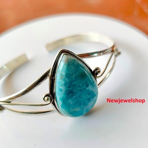 May include: A silver cuff bracelet with a pear-shaped, light blue gemstone. The open-ended bracelet has a delicate design with decorative swirls near the gemstone. The gemstone is set in a silver bezel.