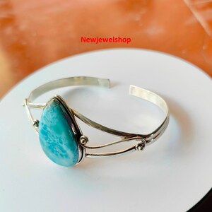 May include: A silver cuff bracelet with a pear-shaped, light blue gemstone. The open-ended bracelet has a double-band design with decorative accents. The gemstone is set in a silver bezel.