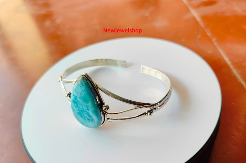 May include: A silver cuff bracelet with a pear-shaped, light blue gemstone. The open-ended bracelet has a double-band design with decorative accents. The gemstone is set in a silver bezel.